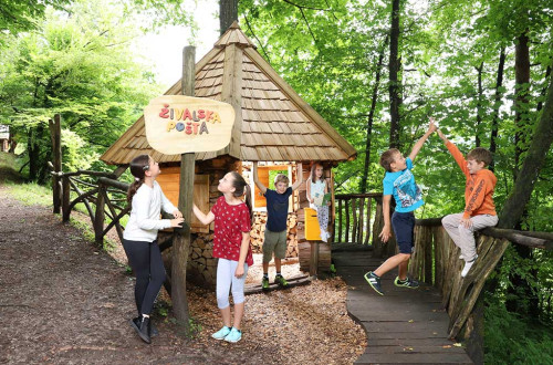 Skupina otrok v Polhovem doživljajskem parku v Polhovem Gradcu v gozdu ob hiški, pred katero je napis Živalska pošta