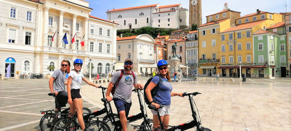 Skupina štirih kolesarjev na Tartinijevem trgu. V ozadju mestna hiša in stavbe.