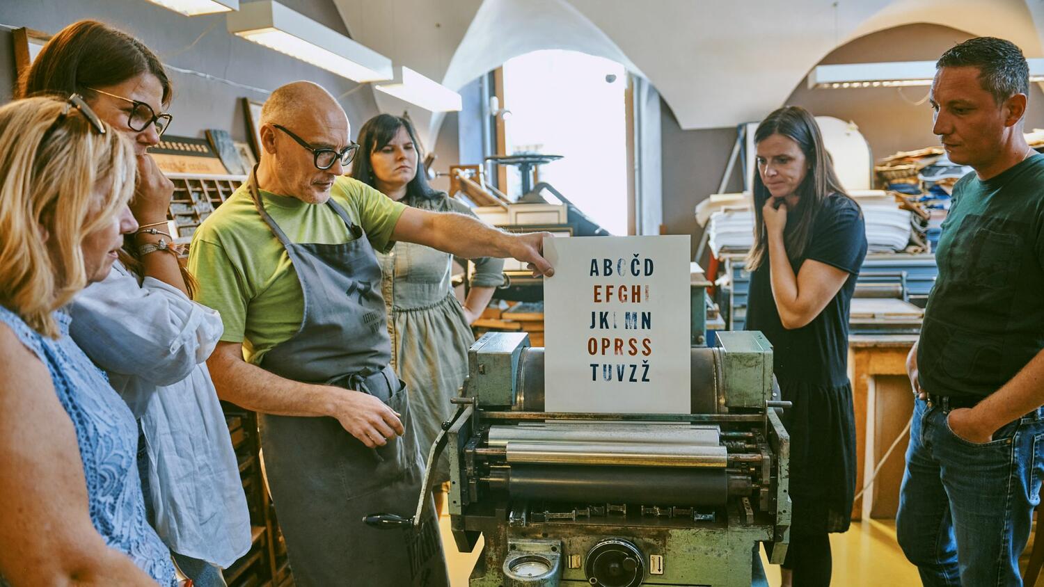 Rokodelska delavnica Natisni svoj plakat v stari tiskarni v Ljubljani – rokodelec kaže skupini pkalat z ročno natisnjeno slovensko abecedo