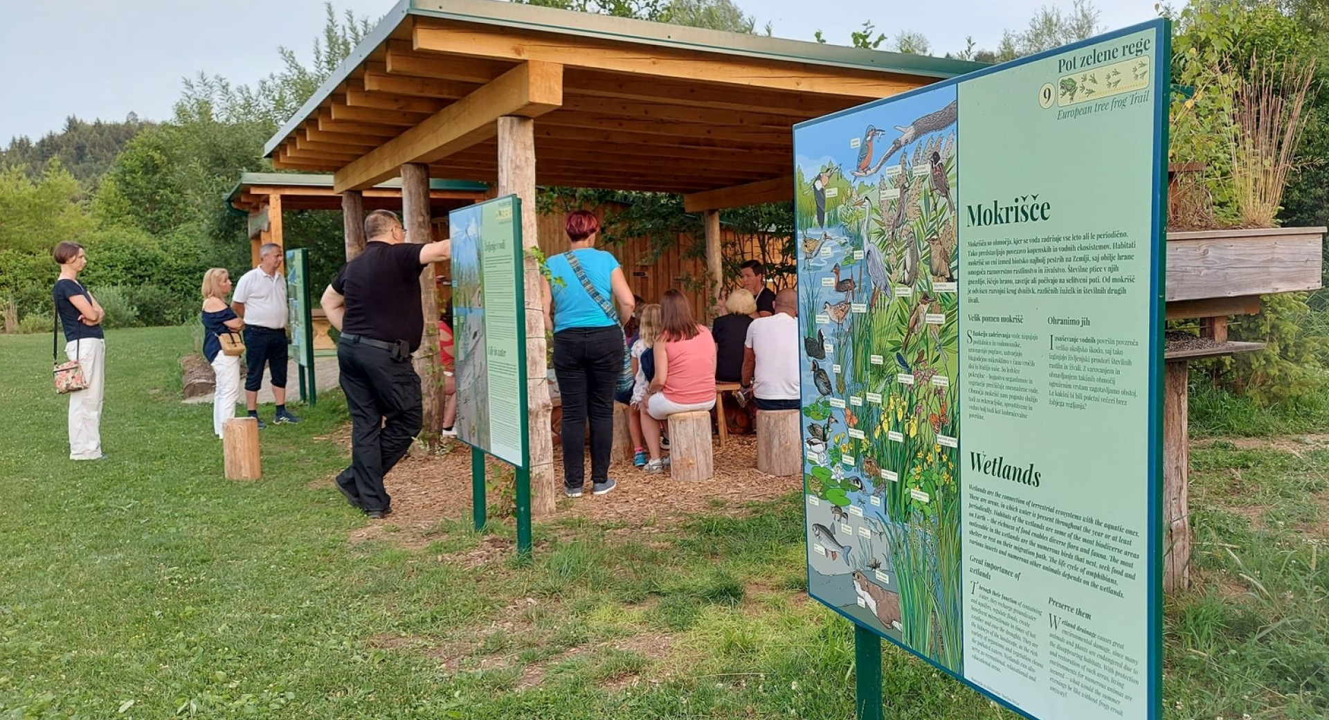 Tematska Pot zelene rege v Vodomčevem gaju v bližini Grosupljega; v ospredju informativna tabla o mokriščih, v ozadju obiskovalci