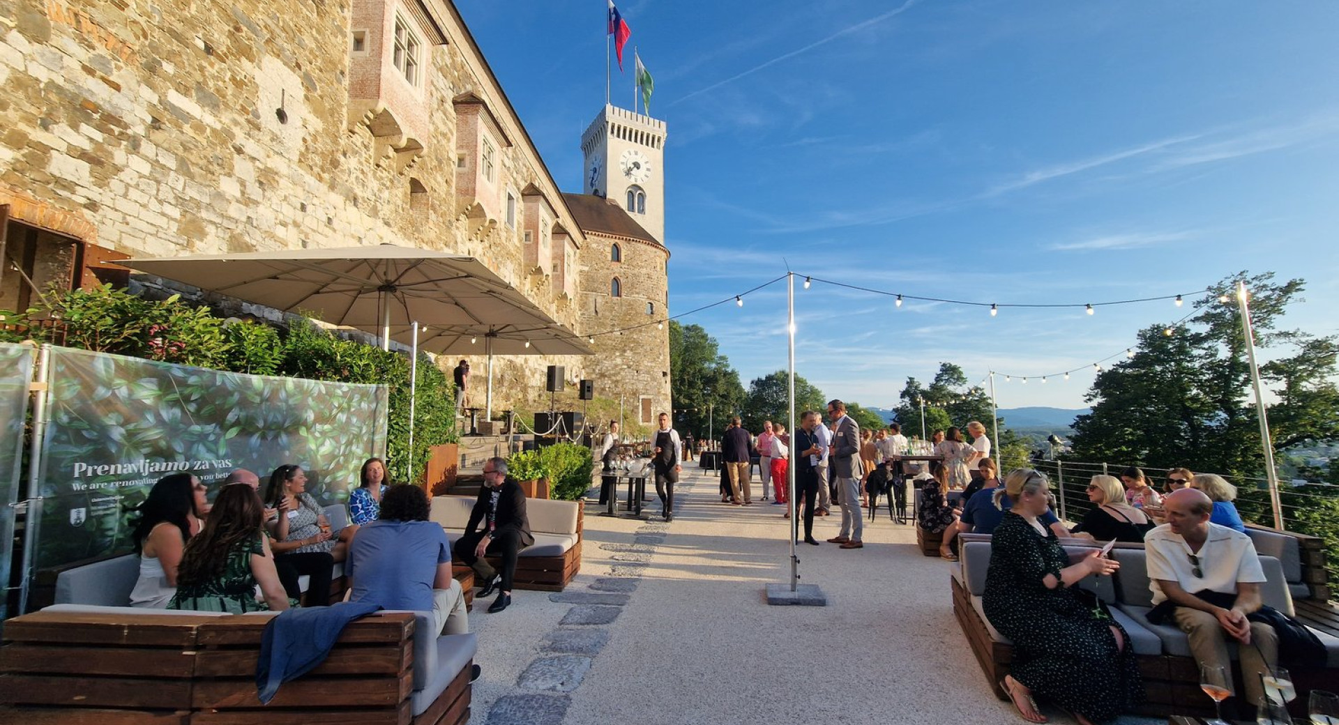 Ljudje sedijo na terasi ob Grajski vinoteki na Ljubljanskem gradu, v ozadju grajski stolp