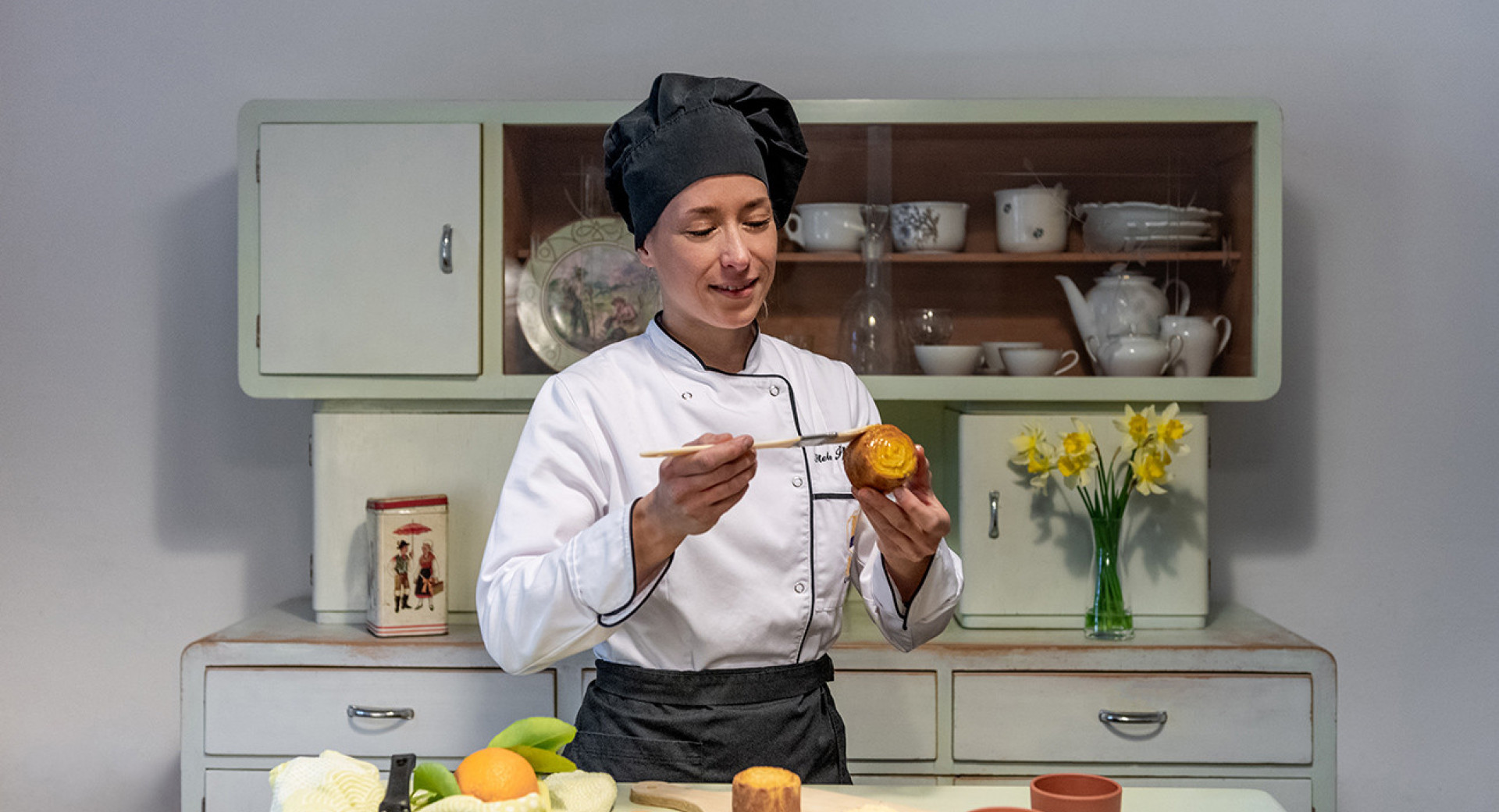 Ženska v kuharskih oblačilih in s kuharsko kapo drži v rokah košček štruklja in ga maže s čopičem, pred njo miza s sestavinami za pripravo končne jedi