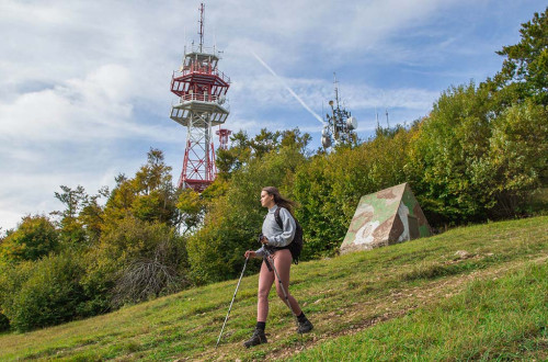 Pohodnica sestopa z vrha hriba. V ozadju nizek pas gozda in antenski stolp.
