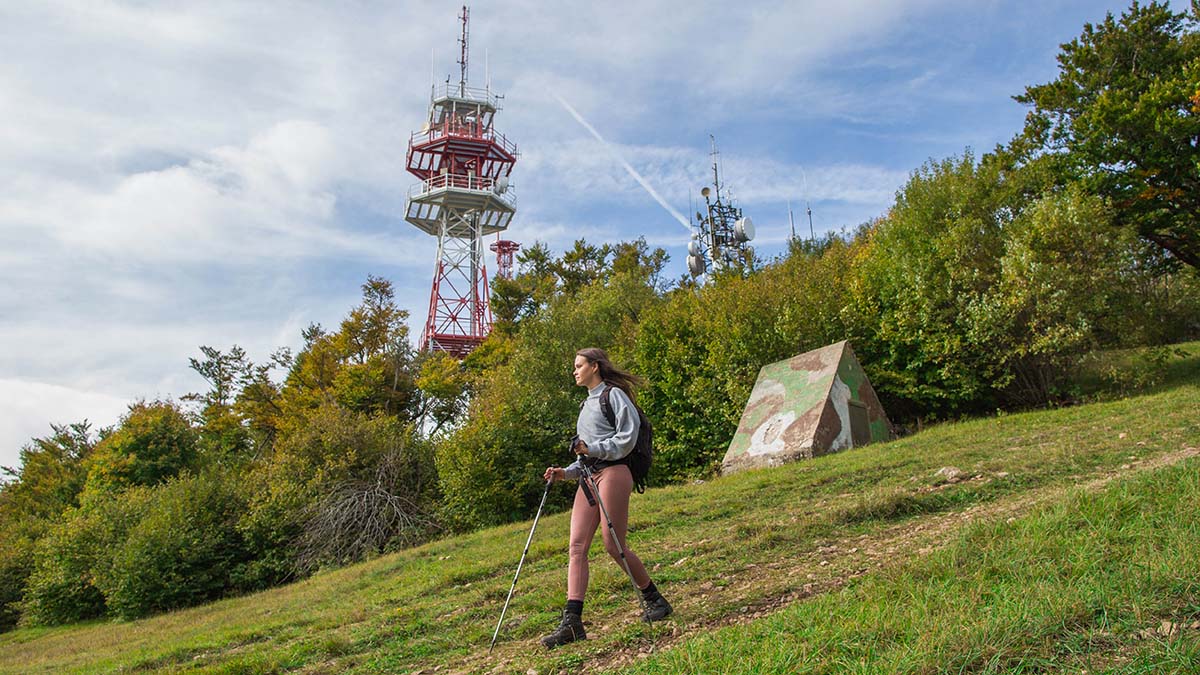 Pohodnica sestopa z vrha hriba. V ozadju nizek pas gozda in antenski stolp.