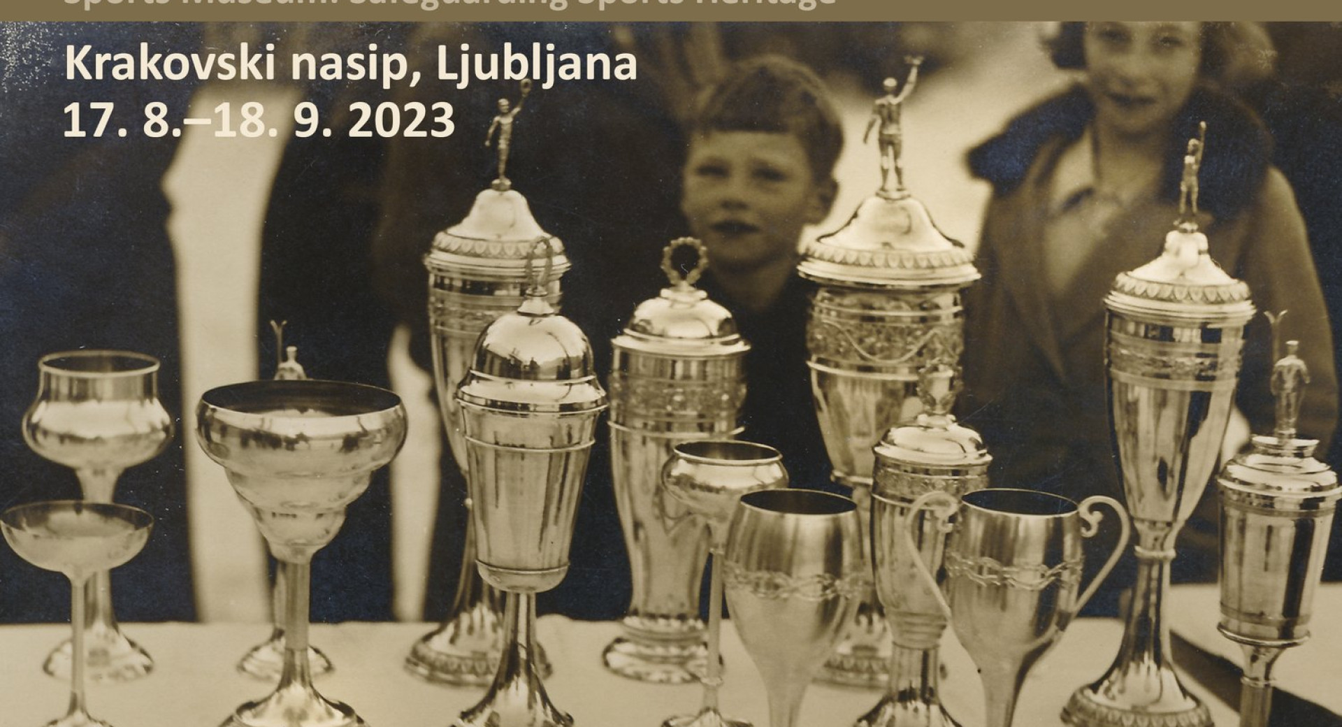 Na crnobeli fotografiji so na mizi pokali. V ozadju otrok in zenska. Gre za napoved razstave Muzej sporta: varuh sportne dediscine.