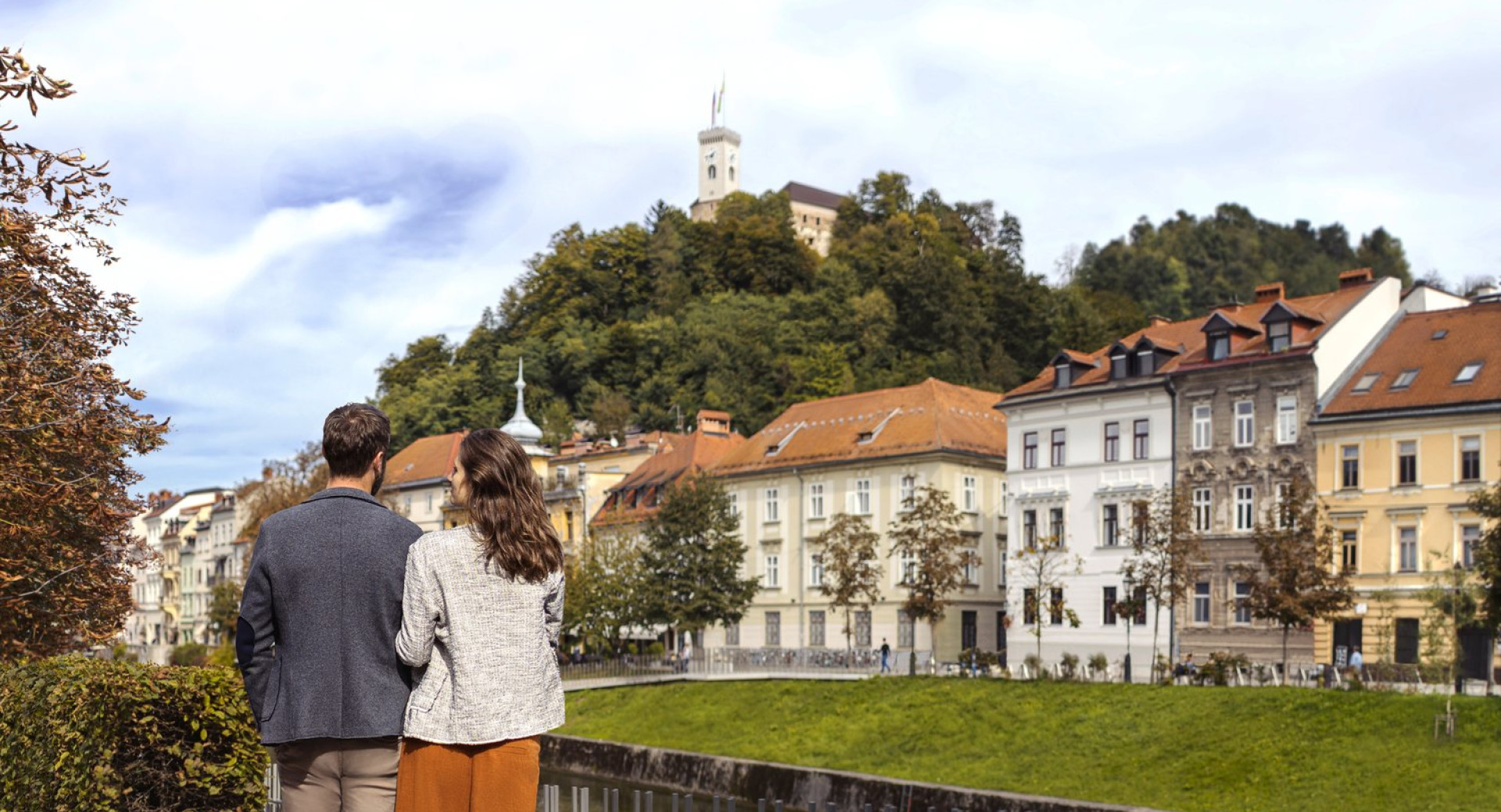 Par stoji ob nabrežju Ljubljanice in zre proti Ljubljanskemu gradu