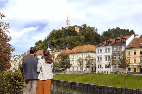 Par stoji ob nabrežju Ljubljanice in zre proti Ljubljanskemu gradu