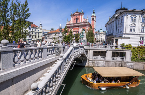 Tromostovje v Ljubljanici, po njem hodijo pešči, v ozadju sta Prešernov trg s spomenikom in Frančiškanska cerkev, po Ljubljanici pa plove turistična ladjica z obiskovalci