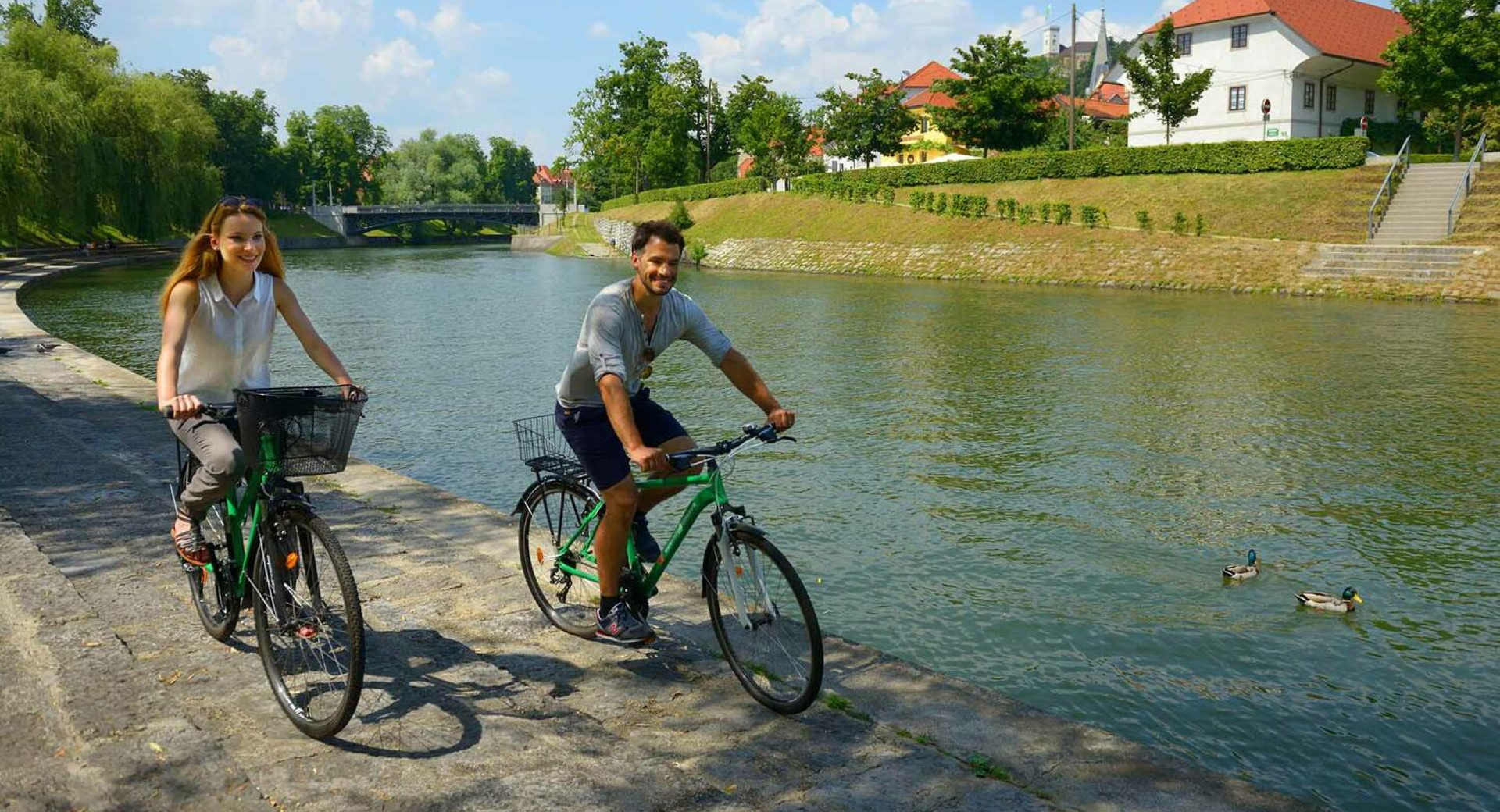 Ženska in moški kolesarita po nabrežju Ljubljanice, v reki rački, v ozadju zelenje in hiše