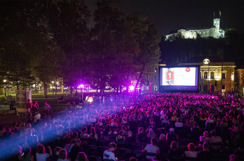Letni kino na trgu v mestu. Obcinstvo na vecerni filmski projekciji, V ozadju Slovenska filharmonija in vise grad.