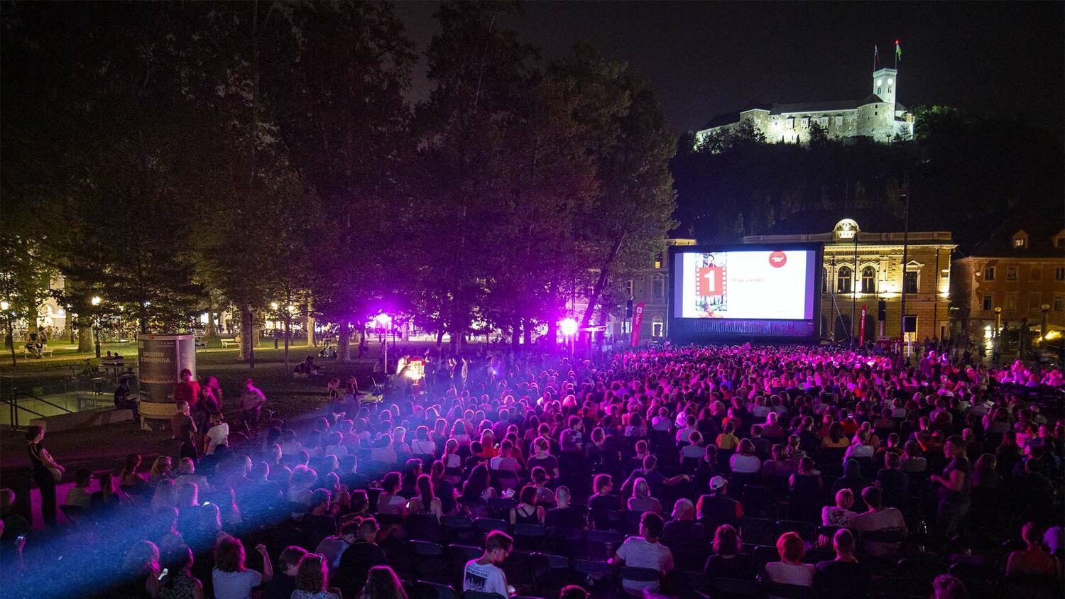 Letni kino na trgu v mestu. Obcinstvo na vecerni filmski projekciji, V ozadju Slovenska filharmonija in vise grad.