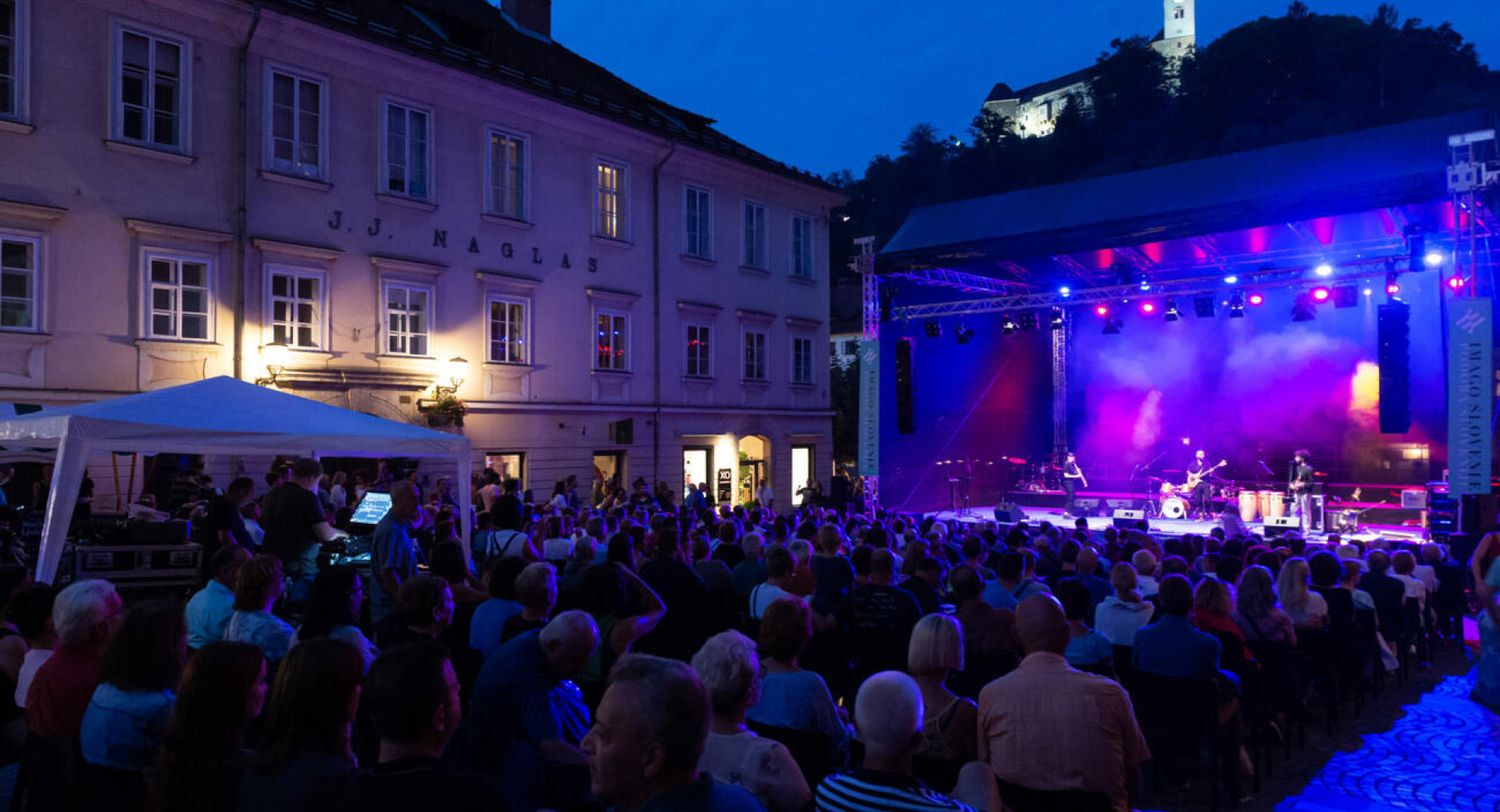Občnstvo na koncertu prireditve Noči v stari Ljubljani, spredaj oder, nad prizoriščem osvetljen Ljubljanski grad