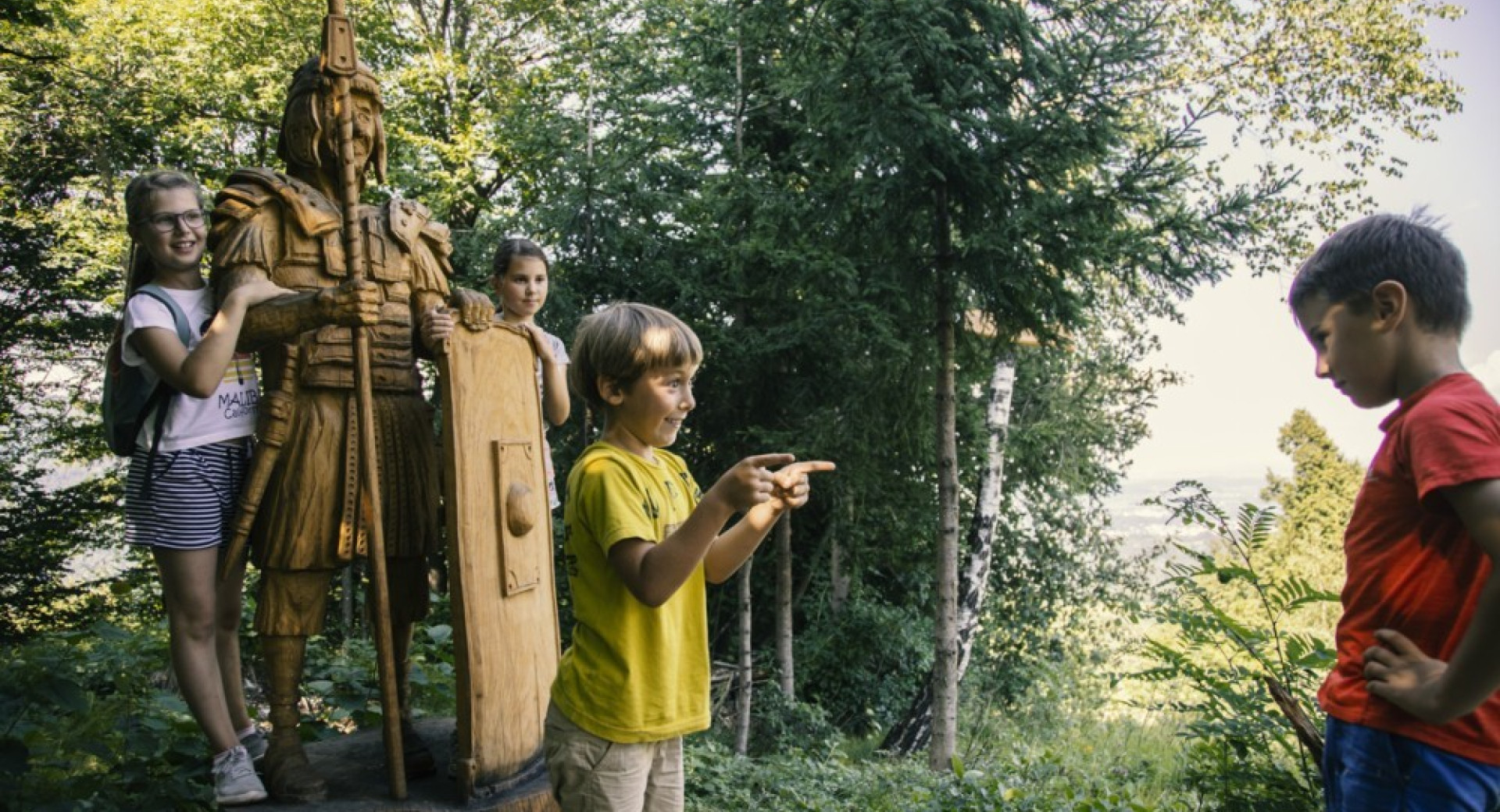 Otroci v gozdu s kipom starodavnega vojaka.