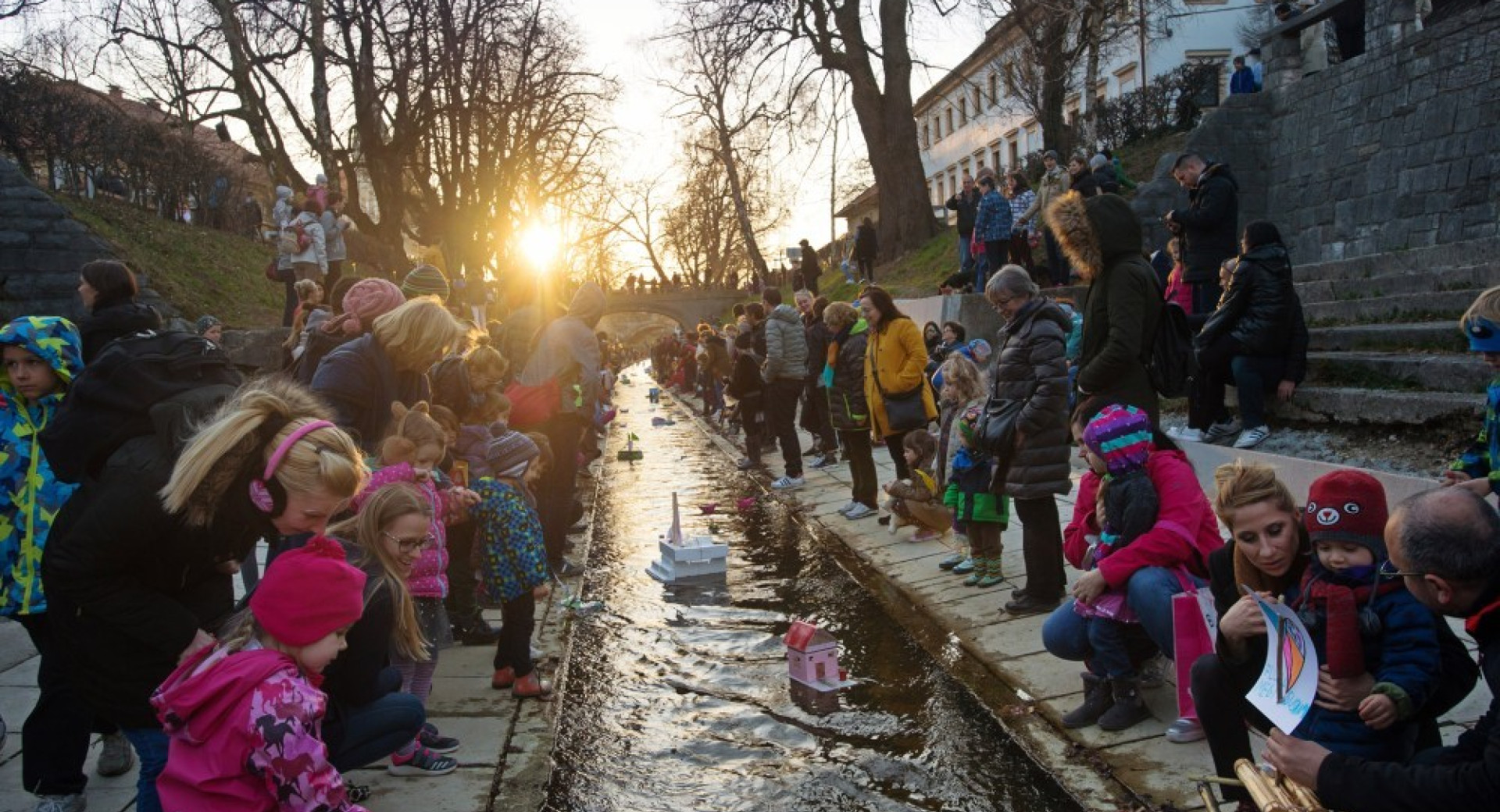 Odrasli in otroci spuscajo papirnate barcice po reki.