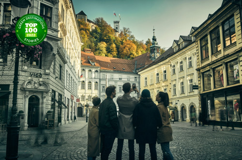 Skupina petih oseb malce pred Mestno hišo v Ljubljani fotografira Ljubljanski grad nad njo