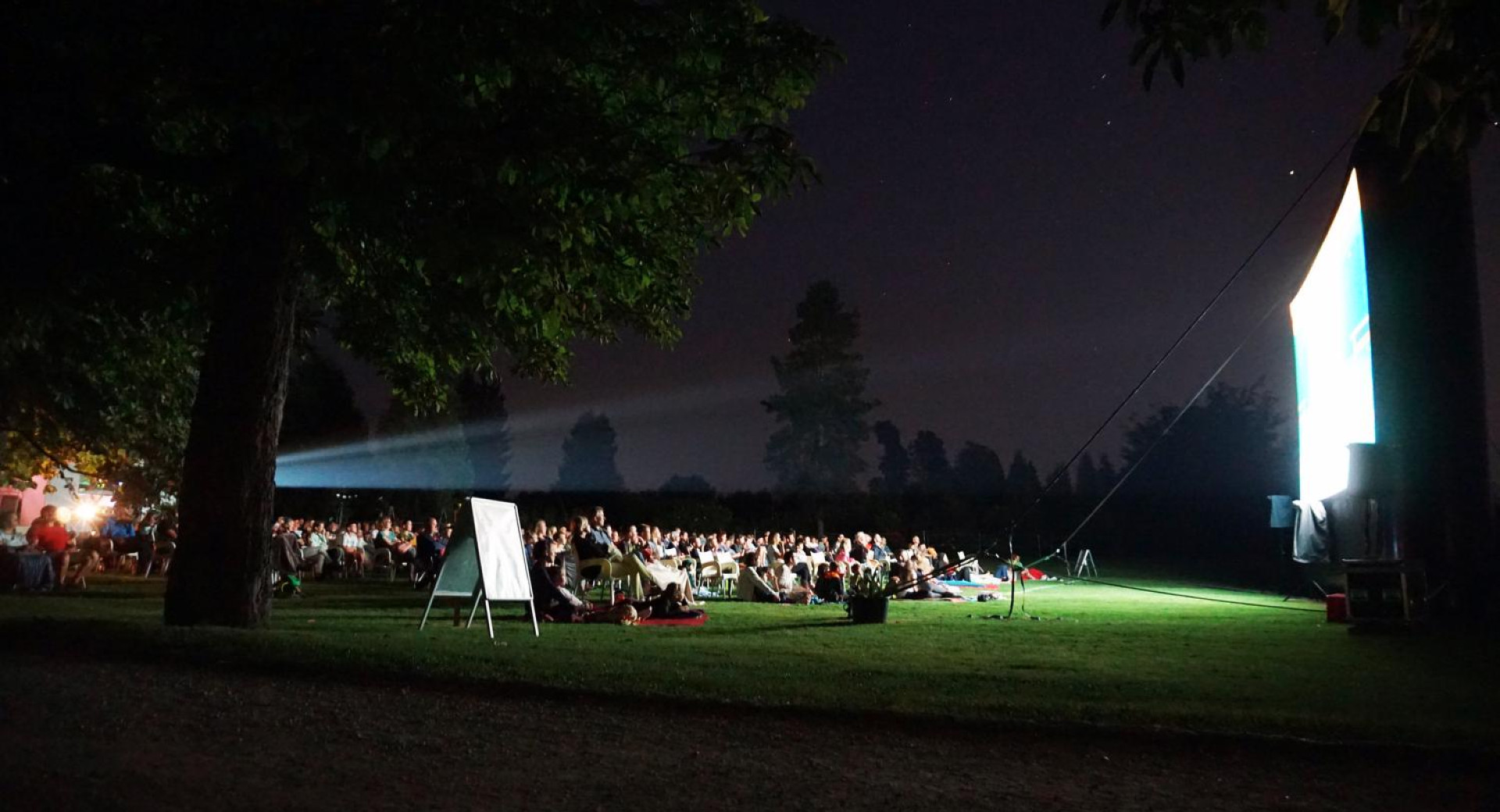 Filmski večer v Arboretumu Volčji Potok; občinstvo sedi v temi na travniku v vrtu.