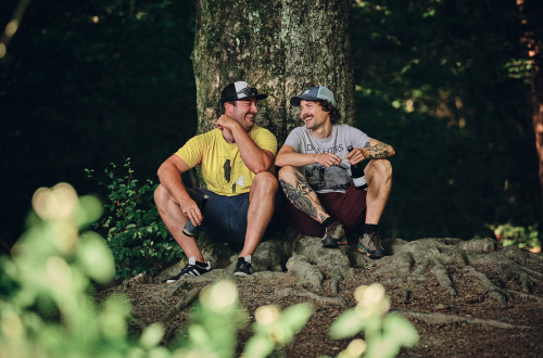 Nasmejan dvojec iz ekipe kamniške pivovarne Barut Brewing and Blending; moška sedita v naravi, ob drevesu
