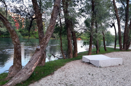 Na fotografiji betonski lezalnik na prodnati plazi v zavetju dreves ob reki