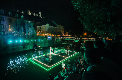 Utrinek z dogodka Odbojka na Ljubljanici; na fotografiji večerni posnetek osvetljenega igrišča na reki med igro, ki jo gledalci spremljajo z mostu in nabrežij