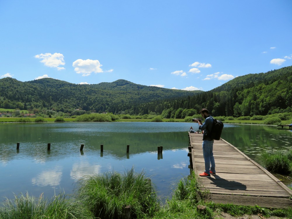 Jezero in fotograf na lesenem pomolu. V ozadju hribi.