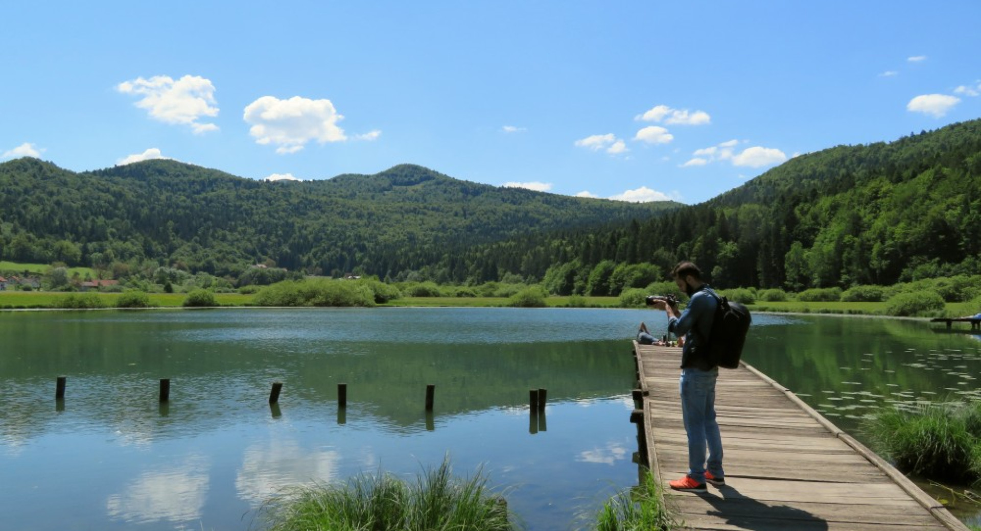 Jezero in fotograf na lesenem pomolu. V ozadju hribi.