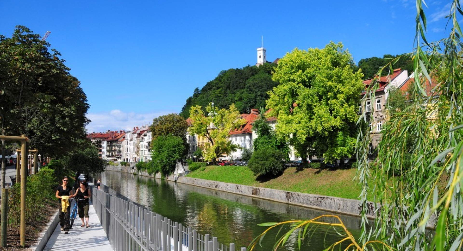 Sprehajalci ob Ljubljanici. V ozadju je Ljubljanski grad.