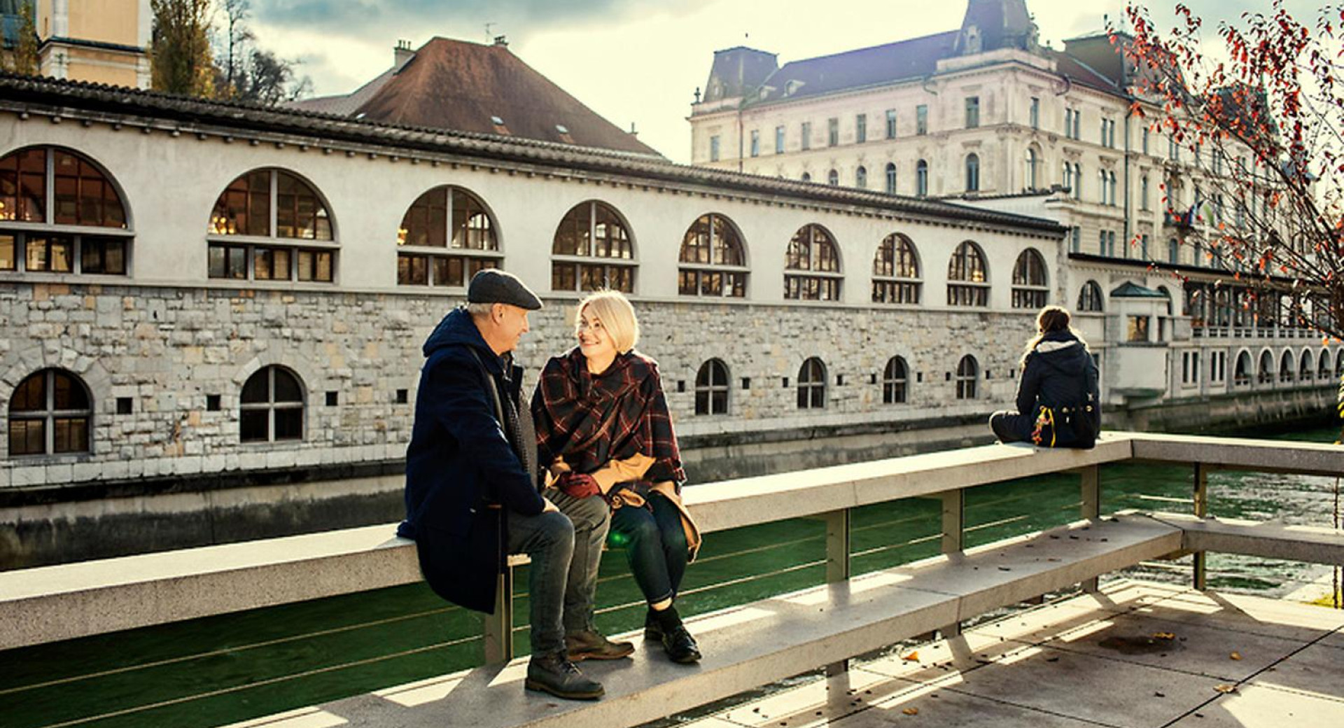Par sedi na klopci ob Ljubljanici. V ozadju Plečnikove tržnice.