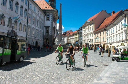 Poletno vzdušje v Stari Ljubljani; v ospredju trije kolesarji pred Robbovim vodnjakom