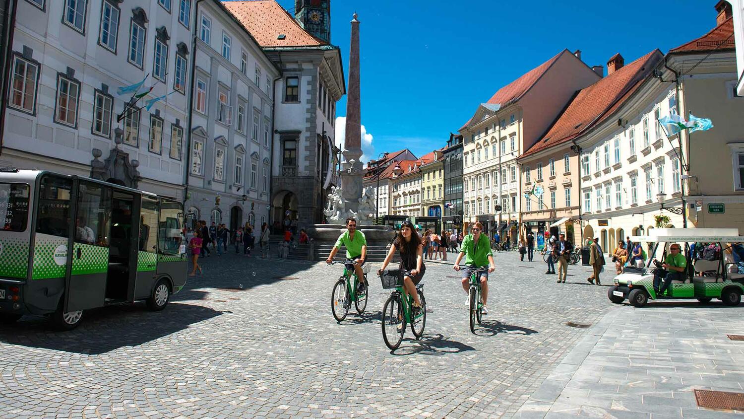 Poletno vzdušje v Stari Ljubljani; v ospredju trije kolesarji pred Robbovim vodnjakom