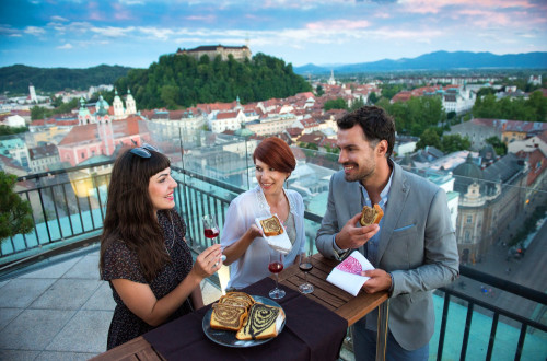Trije prijatelji uživajo ob potici in vinu. V ozadju panorama Ljubljane in Ljubljanski grad.