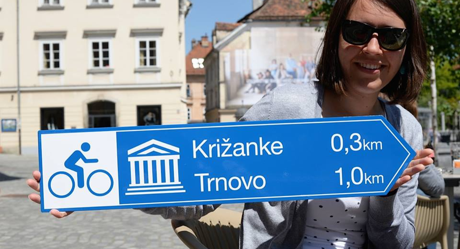 Ženska drži v rokah moder smerokaz za kolesarje z napisoma Križanke in Trnovo (Ljubljana)