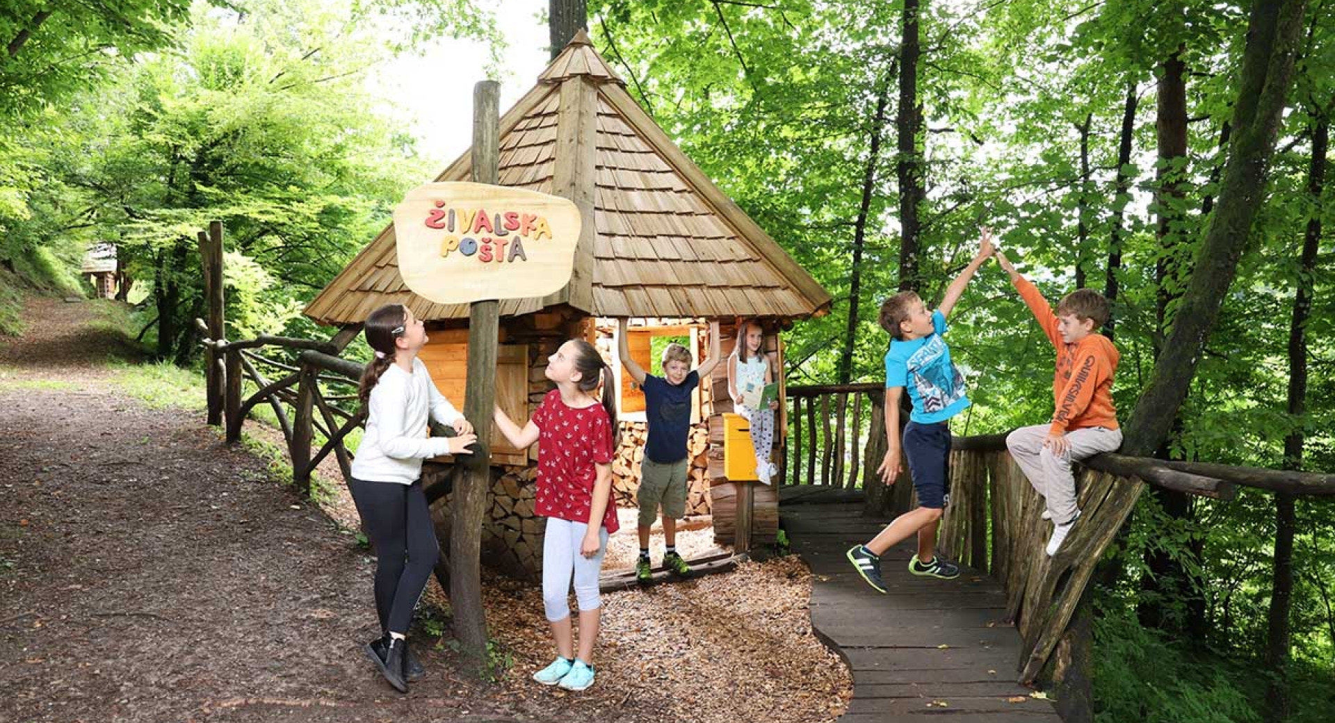 Skupina otrok v Polhovem doživljajskem parku v Polhovem Gradcu v gozdu ob hiški, pred katero je napis Živalska pošta