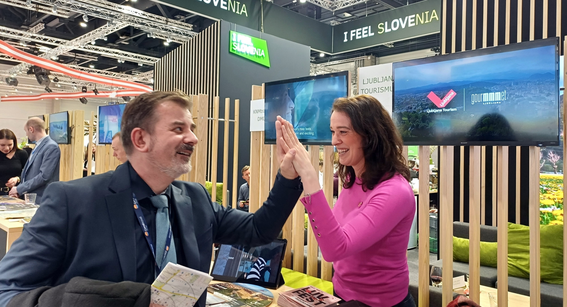 Nasmejana zenska in moski sta udarila z dlanmi nad mizo s promocijskim gradivom. Stojnica z napisom 'I feel Slovenia' v veliki odprti dvorani.