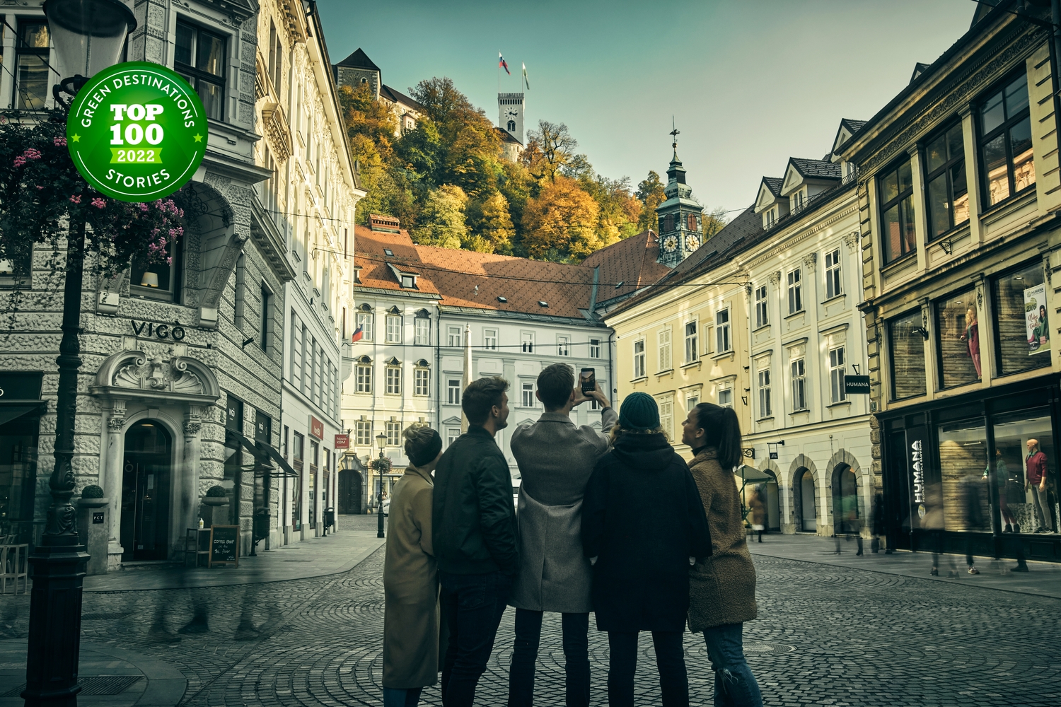 Skupinica ljudi v Ljubljani, zrejo proti Ljubljanskemu gradu; na fotografiji znak uvrstitve na seznam 100 najbolj trajnostnih destinacij na svetu