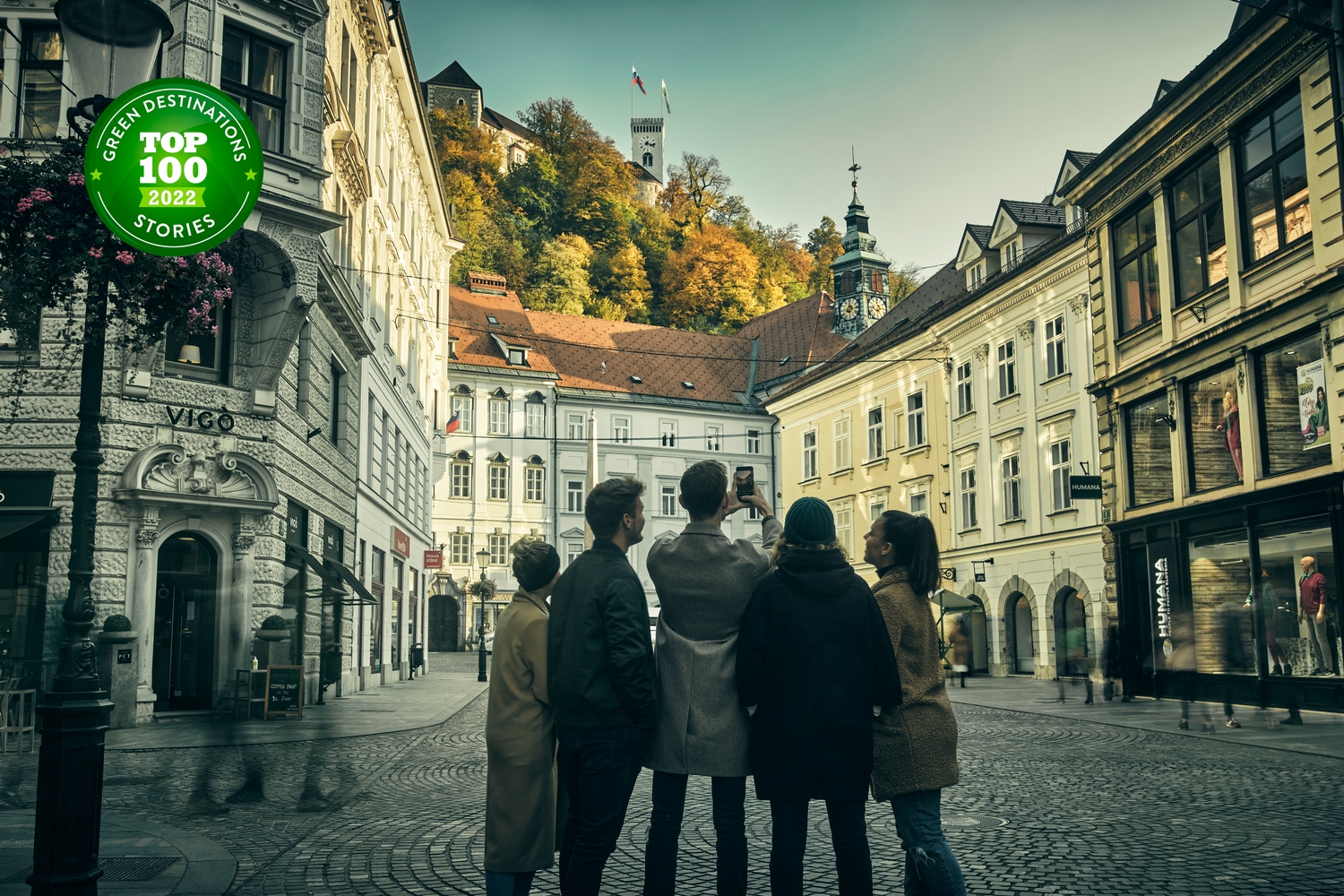 Skupinica ljudi v Ljubljani, zrejo proti Ljubljanskemu gradu; na fotografiji znak uvrstitve na seznam 100 najbolj trajnostnih destinacij na svetu 