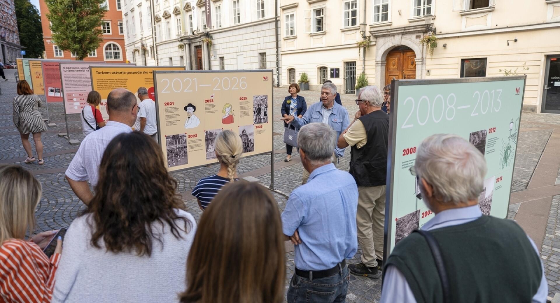 Skupina ljudi si ogleduje informacijske panoje na prostem. V ozadju so vidne zgodovinske stavbe.