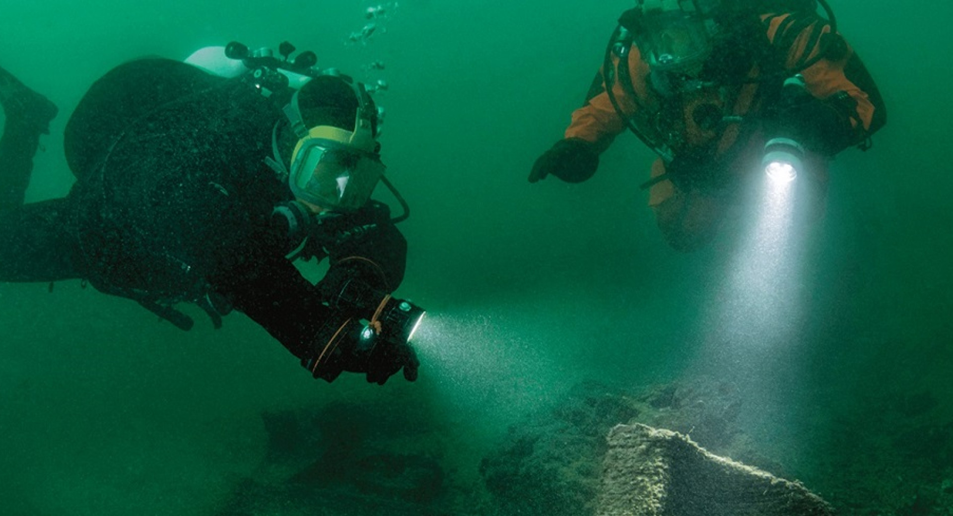 Two divers are exploring the deep water with handheld torches.