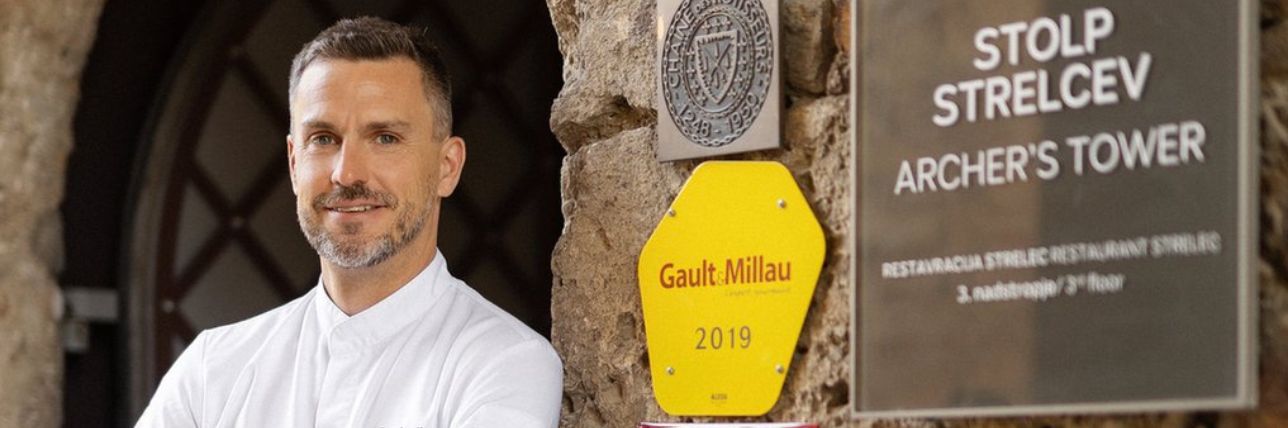 Chef in a white uniform in front of a stone wall with awards hanging on it