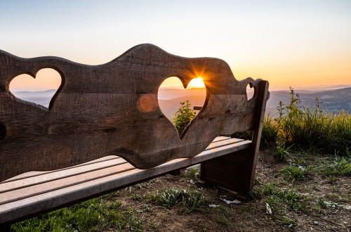 Lesena klopica z izrezanimi srcki v naslonjalu, skozi katere prodirajo soncni zarki. S klopice se odpira razgled na hribe v daljavi, nad katerimi zahaja sonce in nebo zari v toplih zlatorumenih odtenkih.