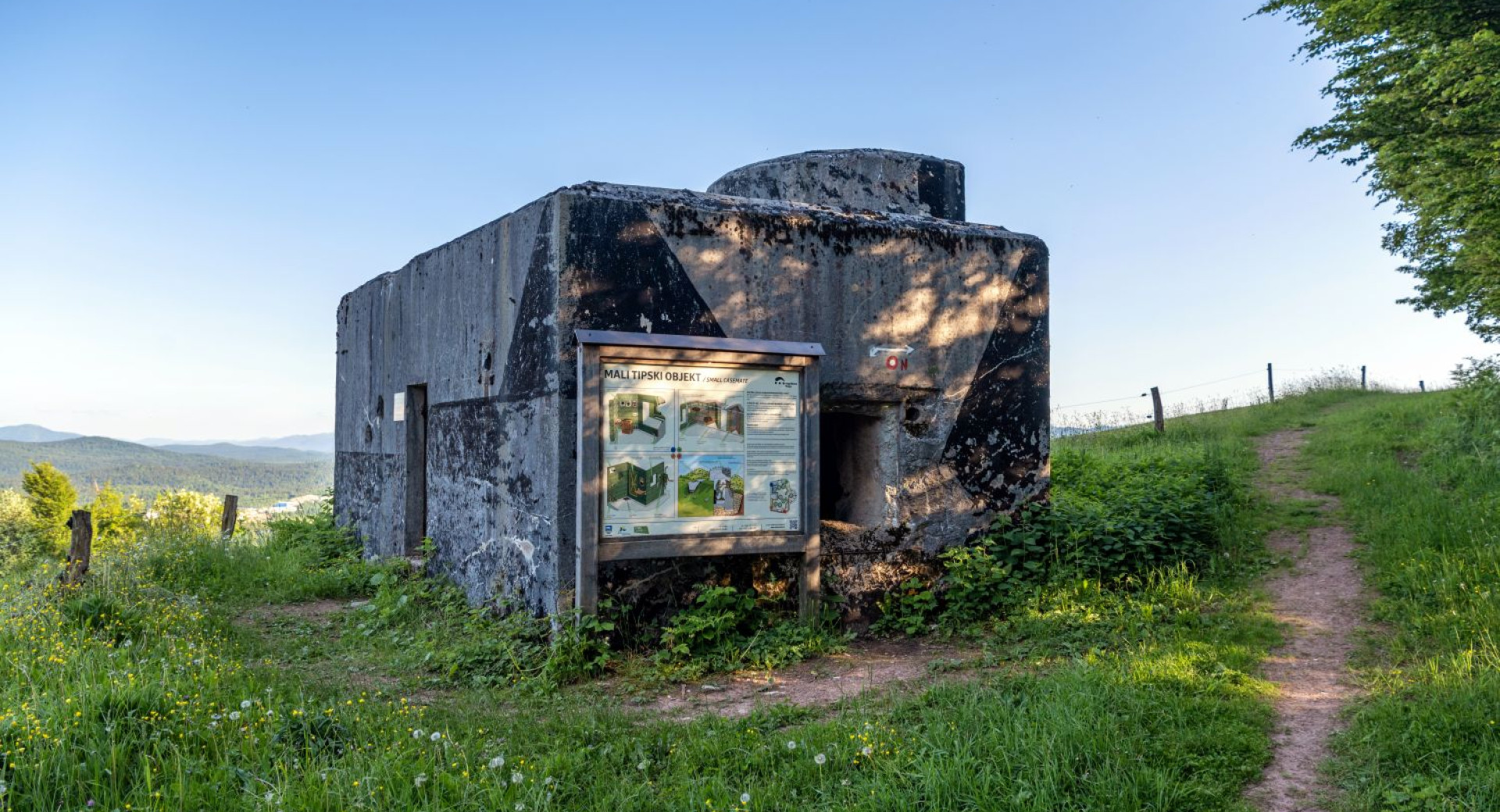 Mali tipski objekt na poti, obkrozen z zeleno travo, z jasnim nebom v ozadju in okoliskih naravnih povrsin.