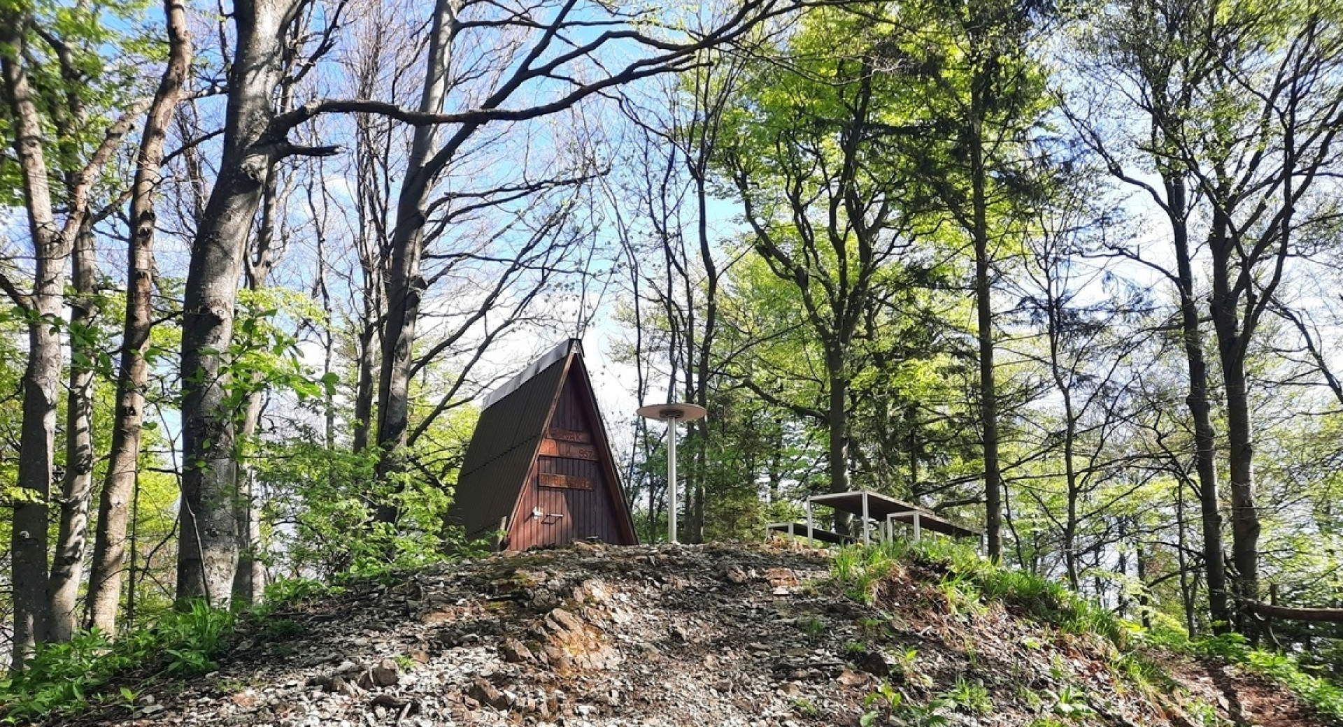 Na vrhu hriba, obdana z gozdom, stoji lesena koča,ob njej pa so postavljene klopice za počitek.
