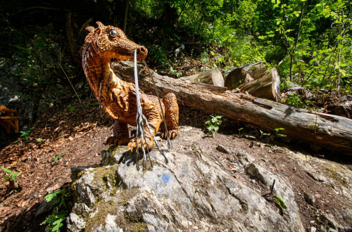 An artificial dragon sits on a rock in the middle of the forest, next to a fallen tree trunk.