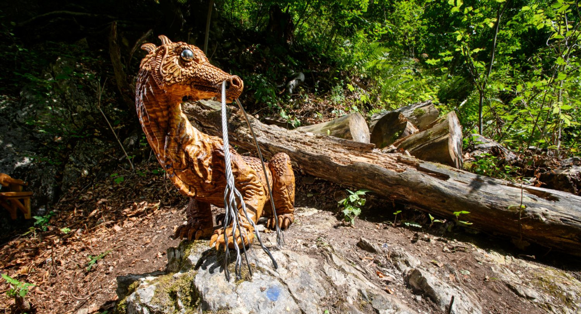 An artificial dragon sits on a rock in the middle of the forest, next to a fallen tree trunk.