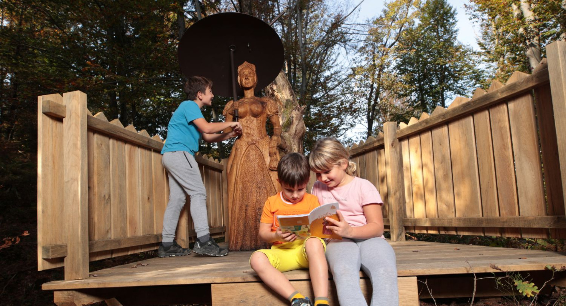 Three children in the forest on a wooden platform are observing the statue of the Cursed Countess and reading a book.