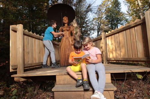 Three children in the forest on a wooden platform are observing the statue of the Cursed Countess and reading a book.