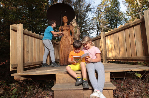 Tri otroci v gozdu na leseni podlagi opazujejo kip Zaklete Grofice in berejo knjigo.