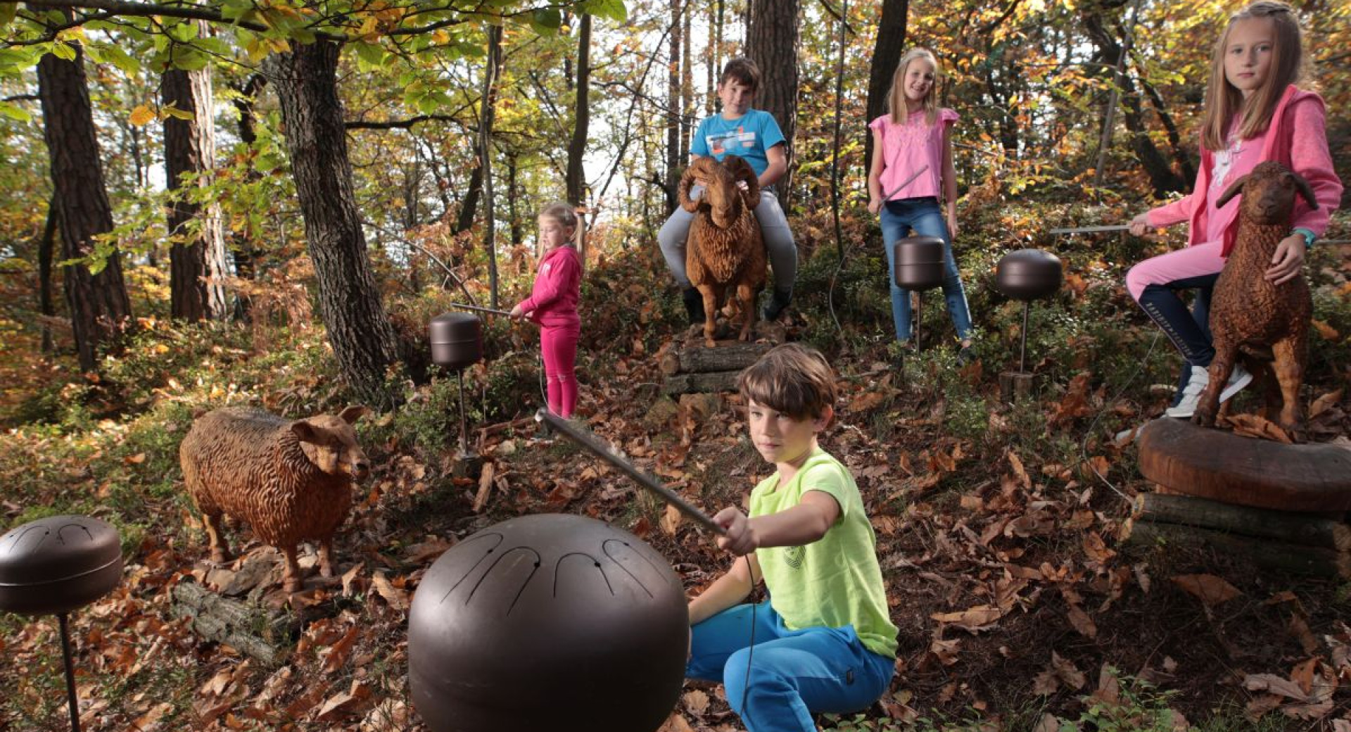 Otroci v gozdu se igrajo z razlicnimi predmeti, medtem ko so okrog njih postavljene lesene ovce.