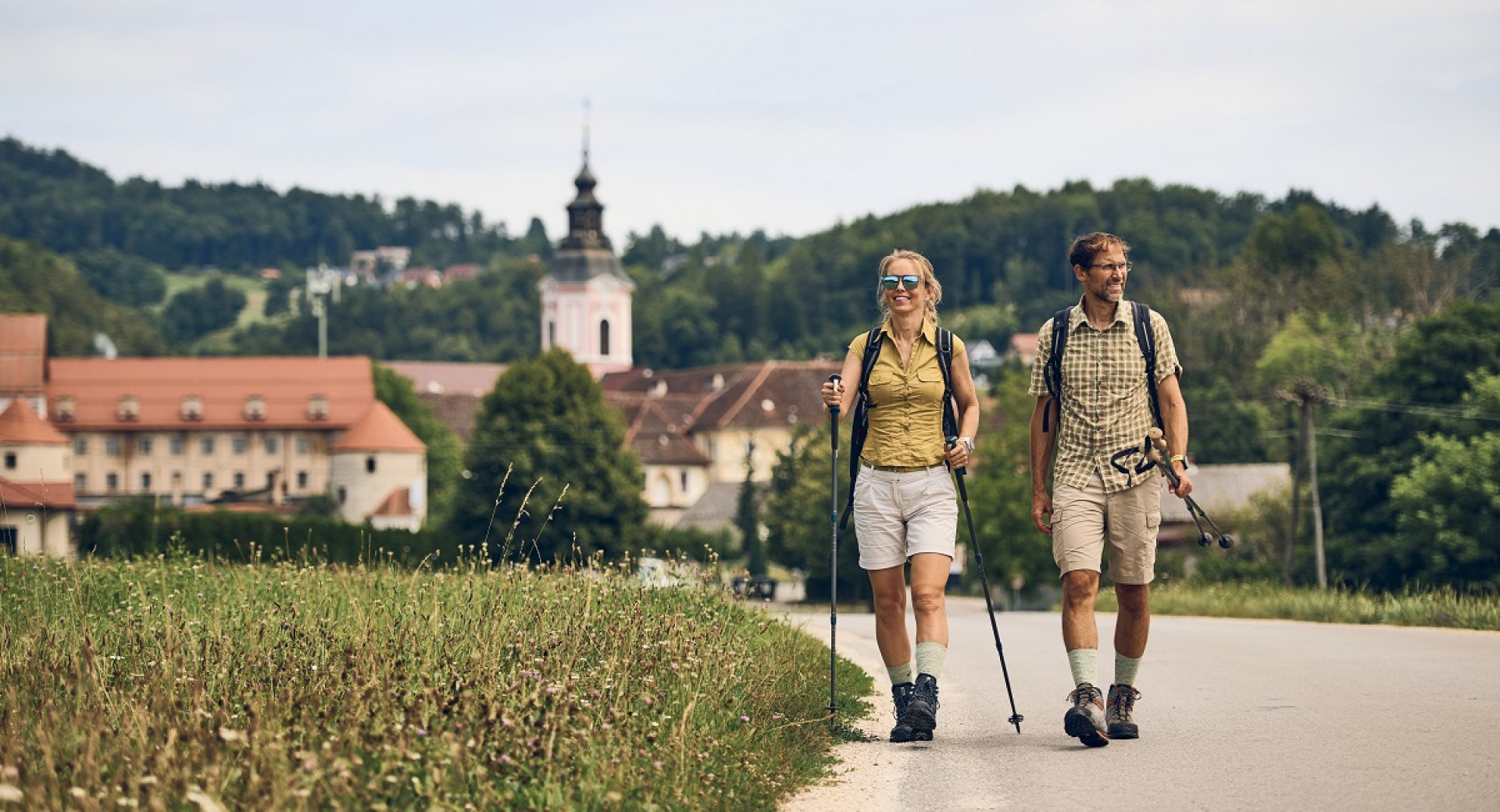 A man and a woman with hiking gear are walking along a road by a meadow, with a settlement in the background featuring a visible church and a hilly landscape.