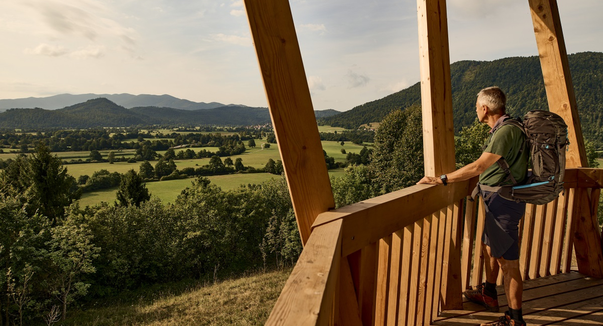 The hiker stands on a wooden observation platform, gazing at the view of the hills and meadows in the valley.