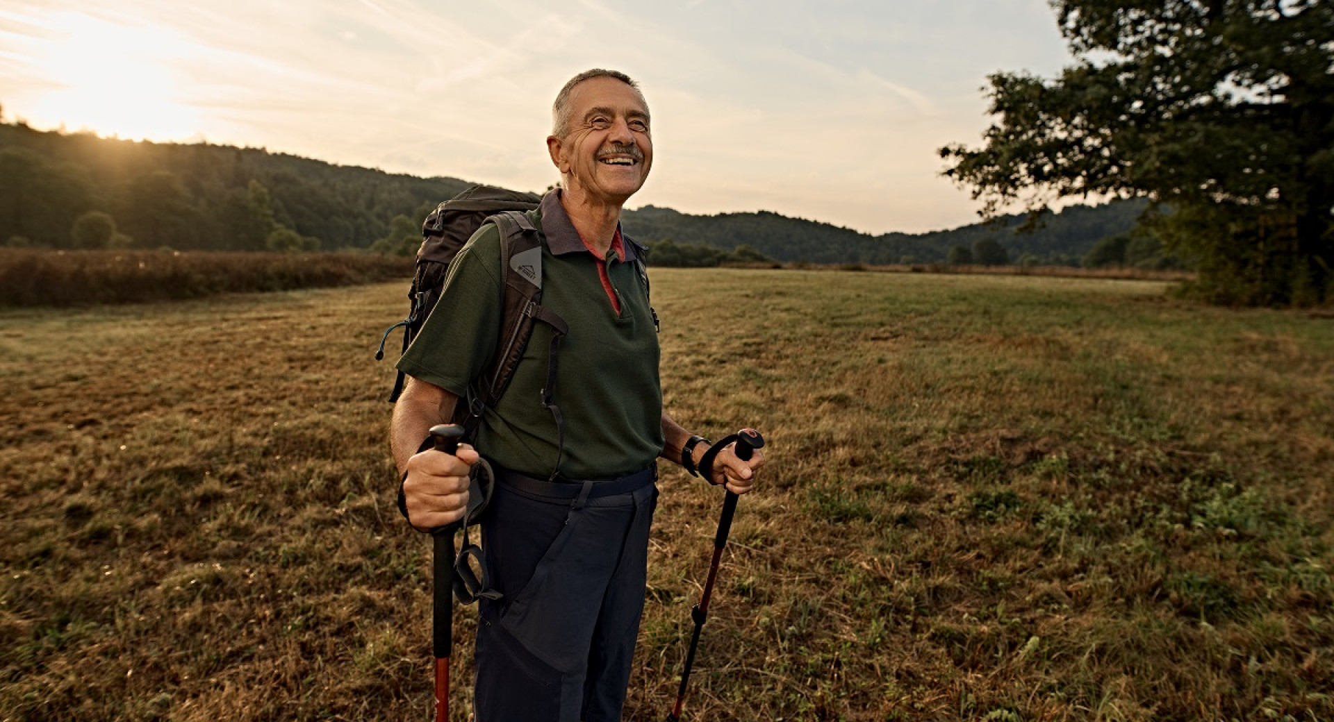 A man is standing on a meadow with a backpack and hiking poles, with a view of the mountains in the background.